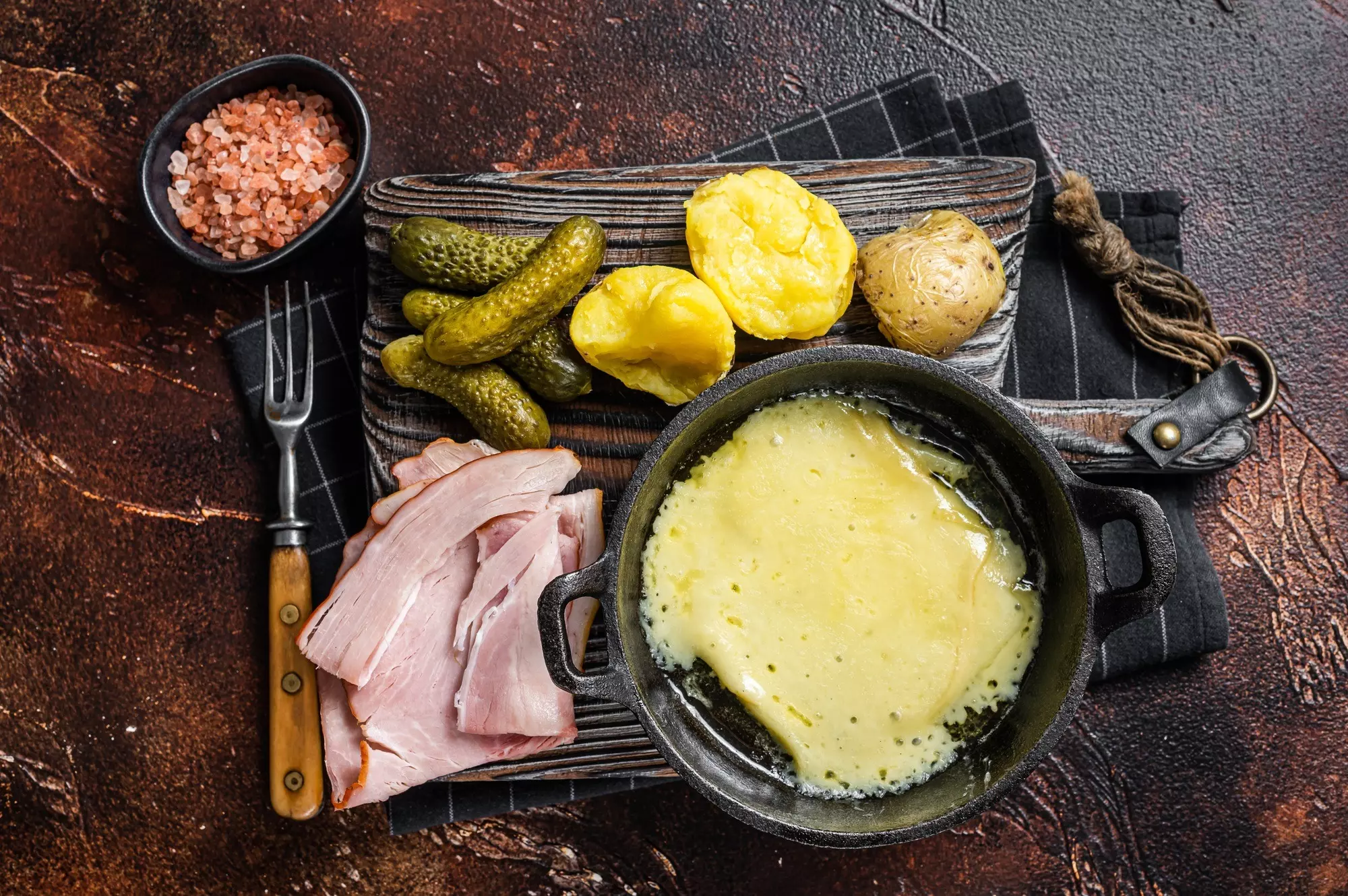 Swiss Raclette Melted cheese with boiled potato and ham on wooden board. Dark background. Top view