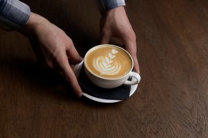 A waiters hands are pictured carefully placing a freshly brewed latte with an intricate foam art des