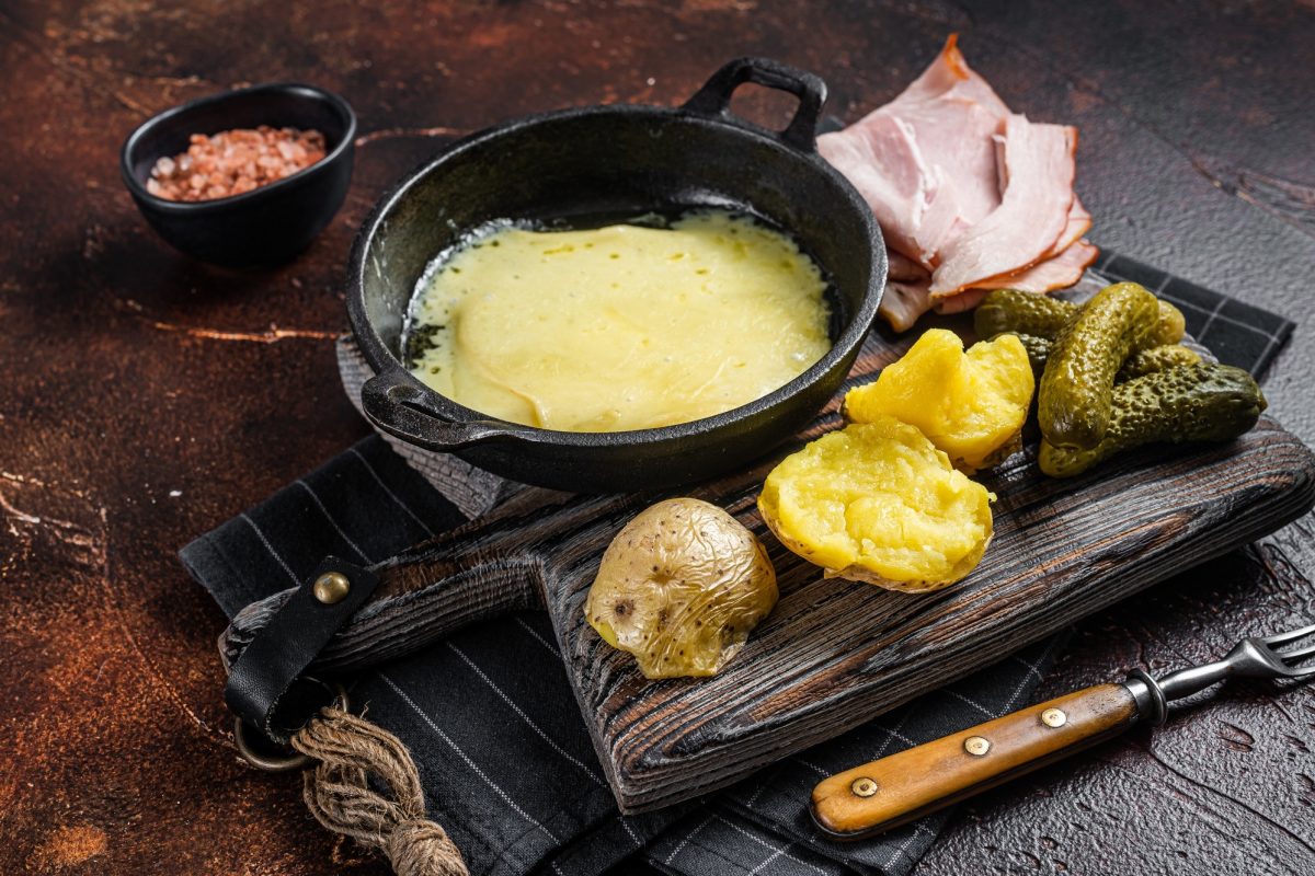 Swiss Raclette Melted cheese with boiled potato and ham on wooden board. Dark background. Top view